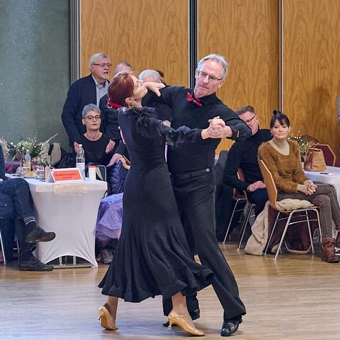 Tanzendes Paar in eleganter Kleidung, umgeben von Zuschauern in einem festlichen Saal mit Holzboden.