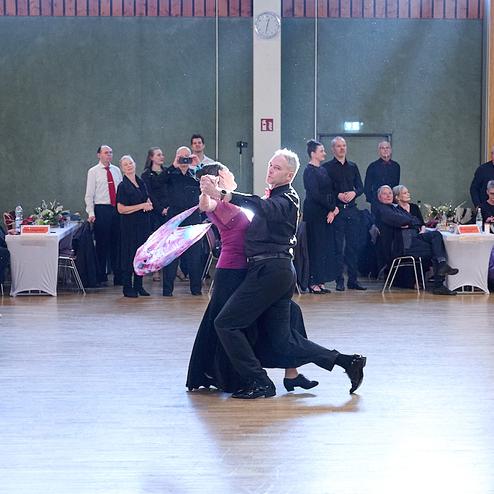 Tanzpaar in eleganter Pose auf der Tanzfläche, Zuschauer in Abendkleidung sitzen an Tischen im Hintergrund.