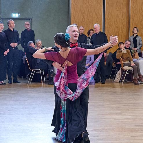 Tanzenden Paar in eleganter Kleidung, umgeben von Zuschauern in einem Tanzsaal mit Holzfußboden.