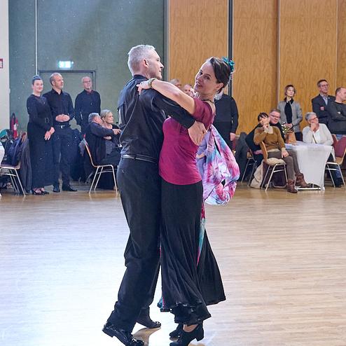 Tanzpaar in eleganter Kleidung tanzt auf einer hellen Tanzfläche, Zuschauer im Hintergrund beobachten aufmerksam.