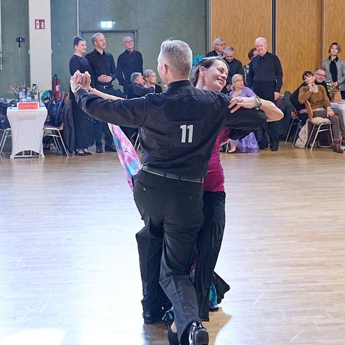 Tanzendes Paar in eleganter Kleidung, umgeben von Zuschauern in einem festlichen Saal.