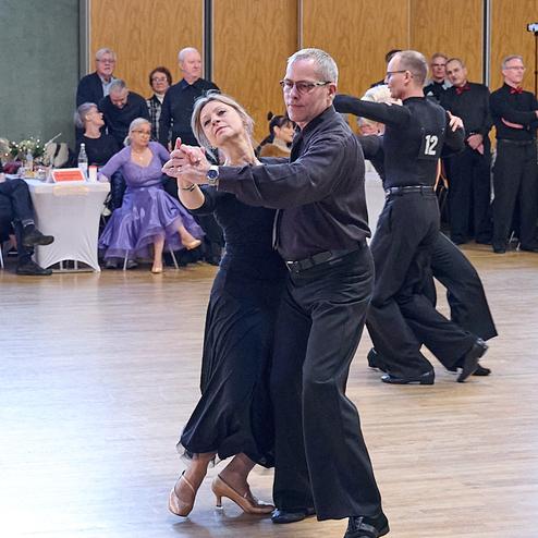Tanzpaar führt einen engagierten Tango auf einer Tanzfläche, Zuschauer beobachten im Hintergrund.