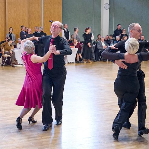 Tanzveranstaltung in einem Saal: Paare tanzen in eleganter Kleidung auf einem Parkettboden, Zuschauer im Hintergrund.