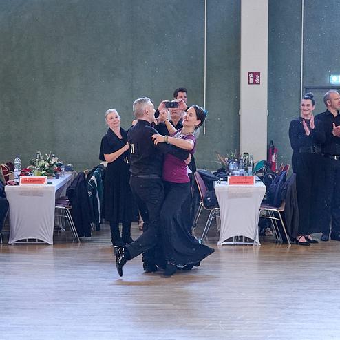 Tanzpaar in eleganter Kleidung tanzt auf einer Veranstaltung, Publikum applaudiert im Hintergrund.