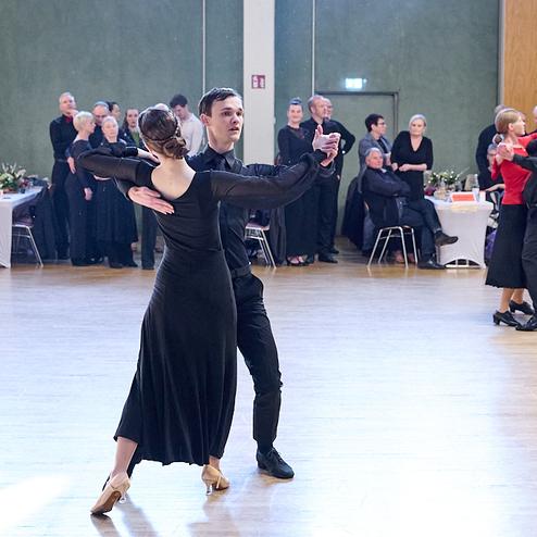 Tanzpaar in eleganter Kleidung tanzt in einem Saal, umgeben von Zuschauern und weiteren Tänzern.