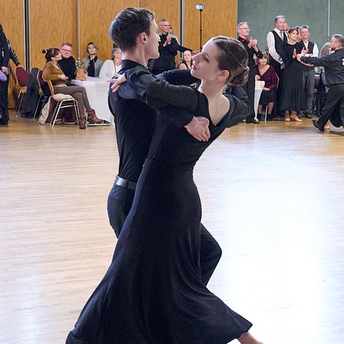 Tanzpaar in eleganter Kleidung führt einen Paartanz in einem festlichen Saal mit applaudierendem Publikum aus.