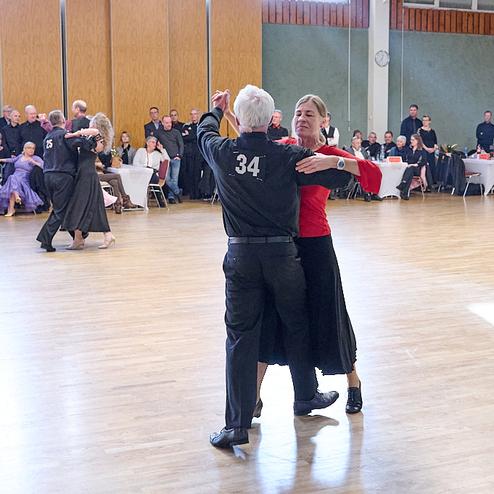 Tanzpaar in eleganter Kleidung tanzt auf einer Holzfläche, Zuschauer sitzen an Tischen im Hintergrund.