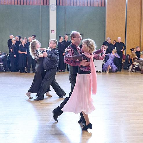 Tanzpaar in eleganter Kleidung tanzt in einem hellen Saal, umgeben von Zuschauern an festlich gedeckten Tischen.