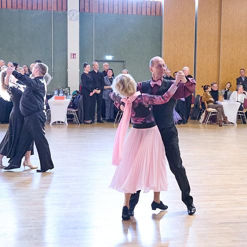 Paare tanzen auf einer Tanzfläche, im Hintergrund Zuschauer an Tischen bei einer Tanzveranstaltung.