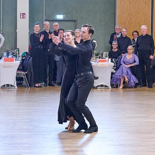 Tanzpaar in eleganter Kleidung tanzt auf einem Parkettboden, umgeben von applaudierendem Publikum.