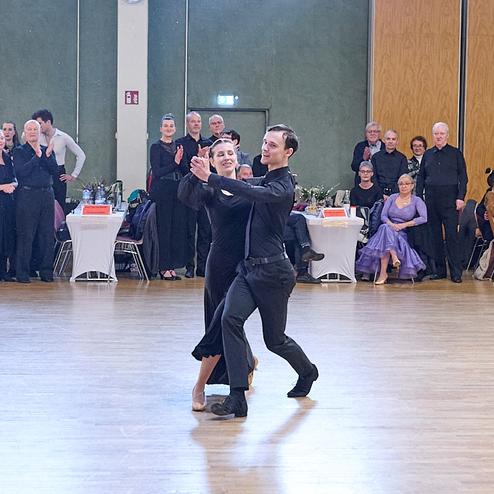 Tanzpaar in eleganter Kleidung tanzt auf einem Parkettboden vor applaudierendem Publikum in einem Saal.