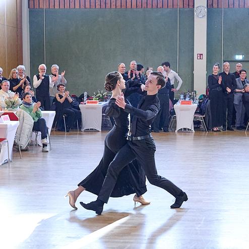 Tanzpaar in eleganter Kleidung führt einen Tanz auf einer Veranstaltung; Publikum applaudiert im Hintergrund.