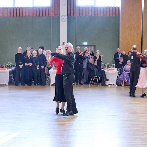 Tanzende Paare in eleganter Kleidung auf einer Tanzfläche, Zuschauer sitzen an Tischen im Hintergrund.