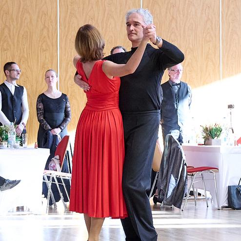 Tanzendes Paar in festlicher Kleidung, Frau im roten Kleid und Mann in schwarzem Oberteil, vor einer tanzenden Menge.