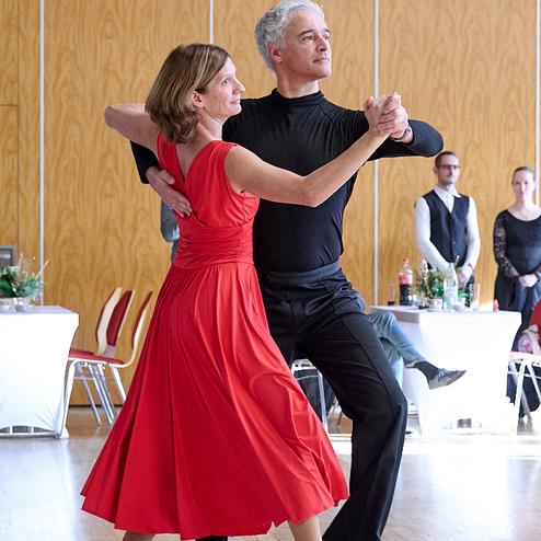 Tanzendes Paar in eleganter Kleidung, der Mann in Schwarz und die Frau im roten Kleid auf einer Tanzfläche.
