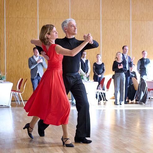 Tanzendes Paar in eleganter Kleidung, umgeben von Zuschauern in einem hellen Saal mit Holzboden.