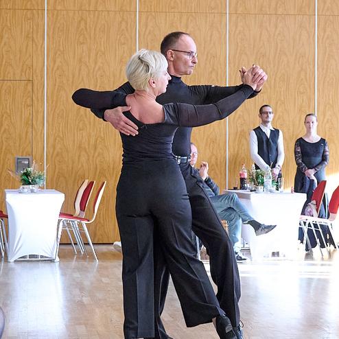 Tanzpaar in eleganter Haltung auf einer Tanzfläche; Zuschauer im Hintergrund beobachten die Darbietung.