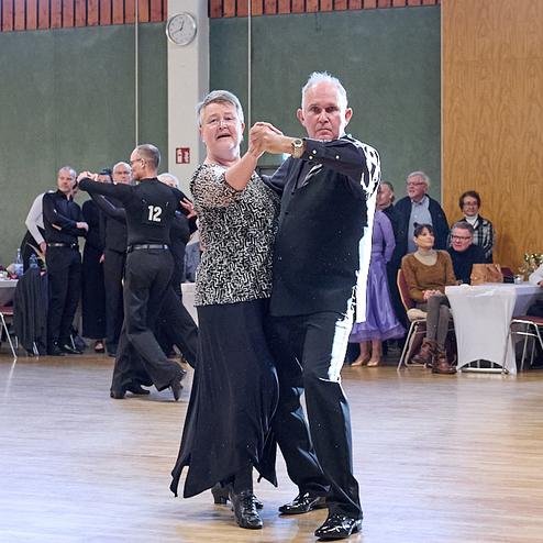 Tanzendes Paar in eleganter Kleidung auf einer Tanzfläche, im Hintergrund weitere Tänzer und Zuschauer.