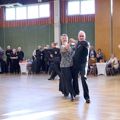 Tanzende Paare in eleganter Kleidung auf einer Veranstaltung, Zuschauer beobachten im Hintergrund.