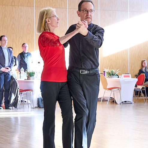 Tanzendes Paar in einem festlichen Saal, gehaltene Hände, Frauen in rotem Oberteil, Mann in schwarzem Hemd.