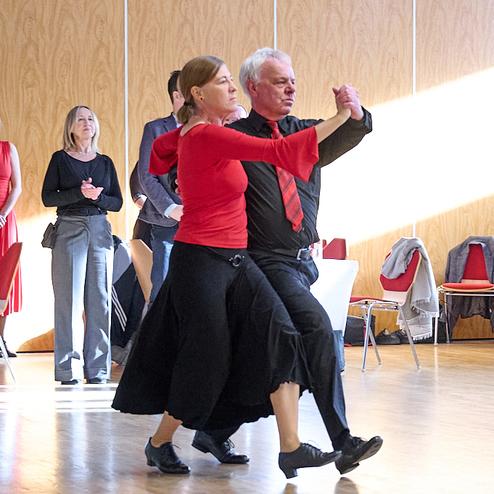Tanzendes Paar in formeller Kleidung, umgeben von Zuschauern in einem hellen Saal mit Holzverkleidung.