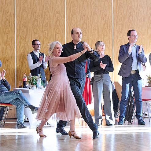 Tanzen eines Paares in eleganter Kleidung, umgeben von applaudierenden Gästen in einem festlichen Raum.