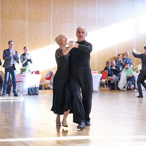 Tanzpaar in eleganter Kleidung tanzt in einer hellen Halle, Zuschauer applaudieren im Hintergrund.