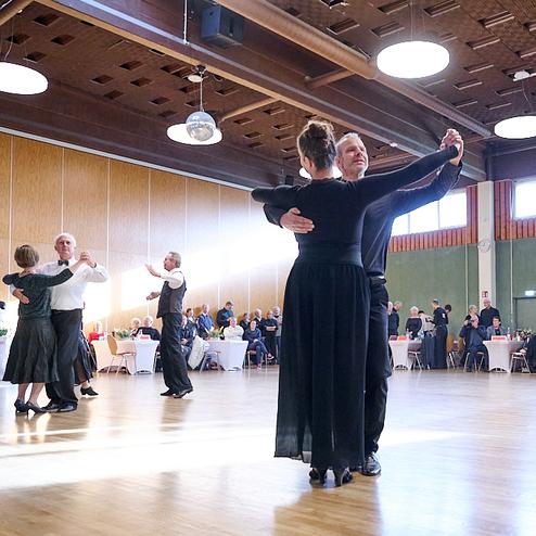 Paare tanzen in eleganter Kleidung auf einer Tanzfläche in einem festlich dekorierten Saal.