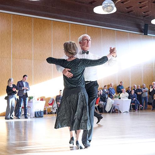 Tanzendes Paar in eleganter Kleidung auf einer Holzfläche, umgeben von Zuschauern bei einer festlichen Veranstaltung.