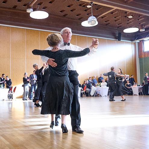 Tanzende Paare in eleganter Kleidung auf einer Tanzfläche in einem festlich dekorierten Raum. Publikum applaudiert.