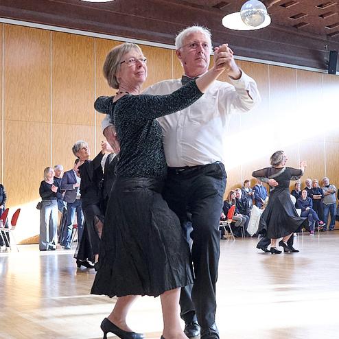 Tanzendes Paar in eleganter Kleidung auf einer Tanzfläche, umgeben von Zuschauern in einem hellen Raum.