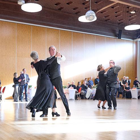Tanzende Paare in eleganter Kleidung auf einer Tanzfläche, umgeben von Gästen an festlich gedeckten Tischen.