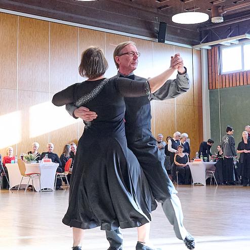 Tanzendes Paar in eleganter Kleidung auf einem Tanzboden, umgeben von anderen Gästen in einem Saal.