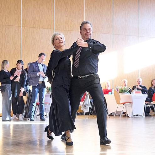 Tanzendes Paar in eleganter Kleidung auf einer Parkettfläche, umgeben von Gästen bei einer Veranstaltung.