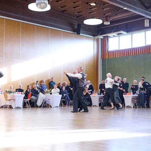 Tanzpaar in eleganter Kleidung beim Senioren-Tanzabend in einem großen Saal mit Zuschauern und festlich gedeckten Tischen.