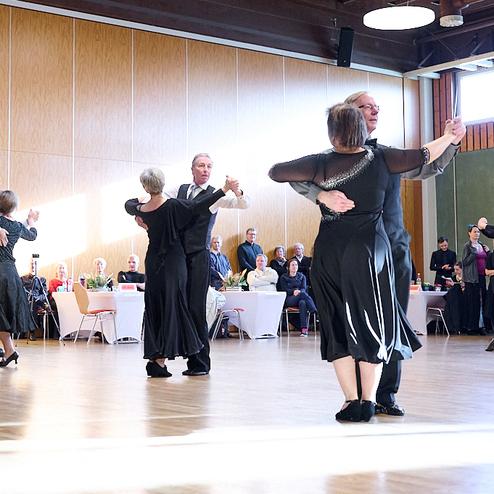 Tanzende Paare in formeller Kleidung auf einer Tanzfläche, mit Gästen im Hintergrund an Tischen.