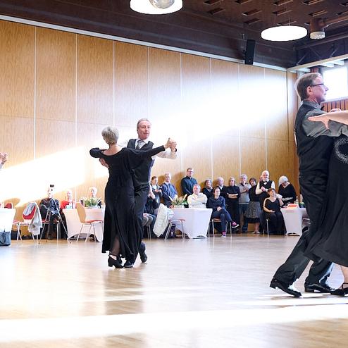 Paare tanzen in eleganter Kleidung auf einer Holzparkettfläche, während Zuschauer in einem Festsaal zuschauen.