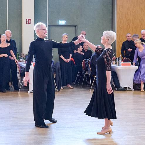 Tänzerpaar in eleganter Kleidung tanzt auf einem Parkettboden, umgeben von Zuschauern bei einer Tanzveranstaltung.