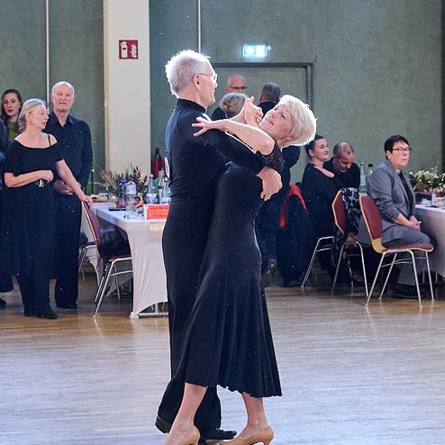 Tanzendes Paar in eleganter Kleidung auf einer Tanzfläche, im Hintergrund Zuschauer und Tischgedecke sichtbar.