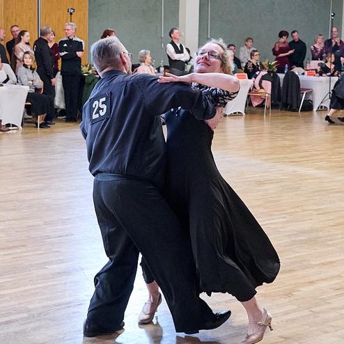 Tanzpaar in eleganter Kleidung führt einen Walzer auf einer Tanzfläche vor einem Publikum aus Gästen vor.