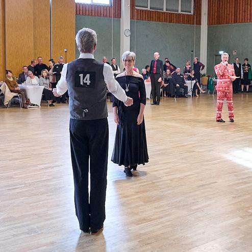 Tanzendende Paare in eleganter Kleidung auf einer Tanzfläche, im Hintergrund Zuschauer an Tischen.