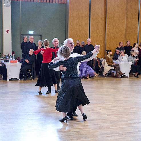 Tanzendes Paar in eleganter Kleidung auf einer Tanzfläche, umgeben von Zuschauern und Tischen im Hintergrund.