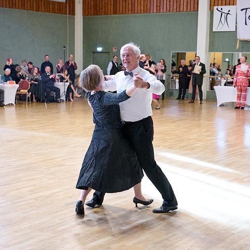 Tanzendes Paar in formeller Kleidung auf einer Tanzfläche, umgeben von Zuschauern und Tischen im Hintergrund.