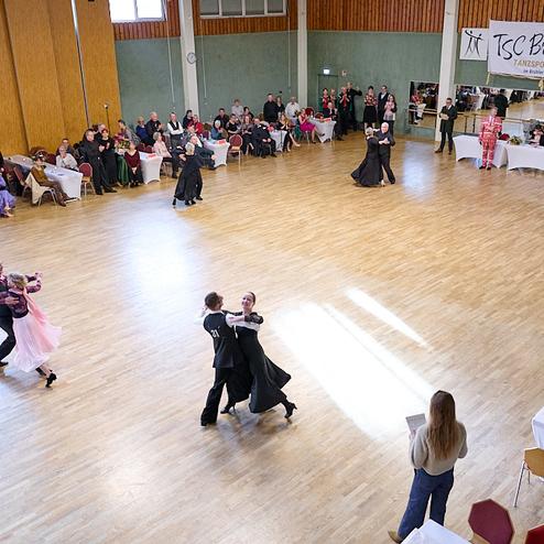Tanzsportturnier im Saal mit tänzerischen Paaren und Publikum an Tischen, im Hintergrund eine Band und eine Banner.