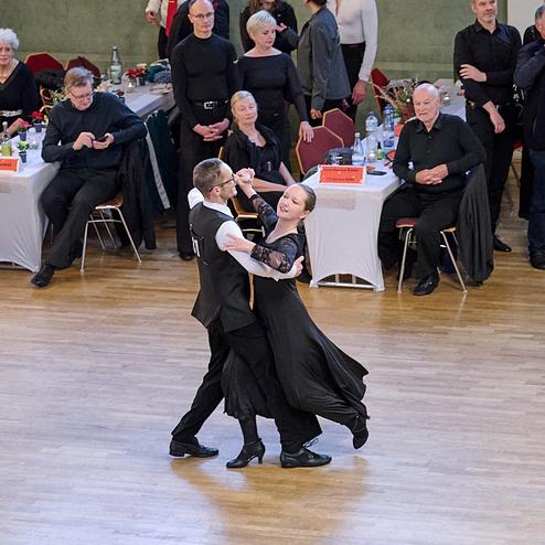 Tanzpaar in eleganter Kleidung tanzt auf einer Holzbühne, während das Publikum aufmerksam zusieht.