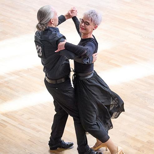 Tanzpaar in eleganter Kleidung, das sich auf einem hellen Parkettboden im Cha-Cha bewegt.