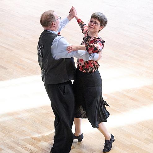 Tanzpaar in eleganter Kleidung, das eng umschlungen auf einem Holzboden tanzt. Helle Lichtstrahlen fallen ein.