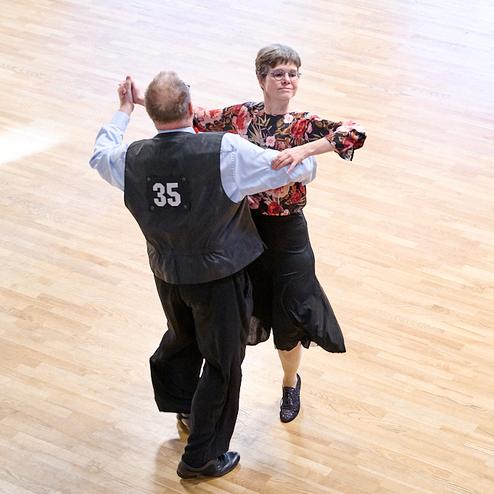 Tanzendes Paar in formeller Kleidung auf einem Holzboden, Mann mit Nummer 35 auf dem Rücken, Frau in bunter Bluse.