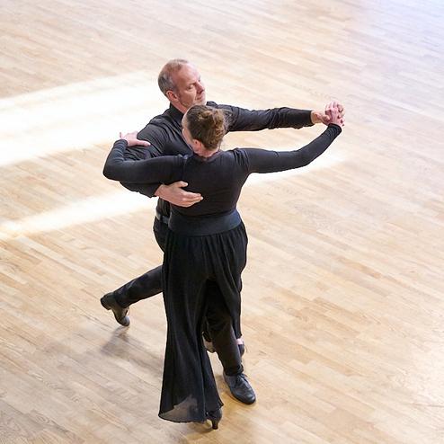 Tanzendes Paar in eleganter Kleidung auf einem Holzfußboden; sie stehen eng umschlungen in einer Tanzpose.