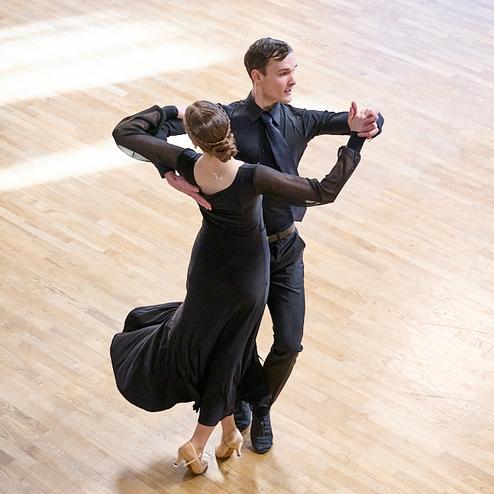 Tanzendes Paar in eleganter Kleidung auf einem hellen Holzboden, der männliche Tänzer führt die Dame in einem Schwarzkleid.